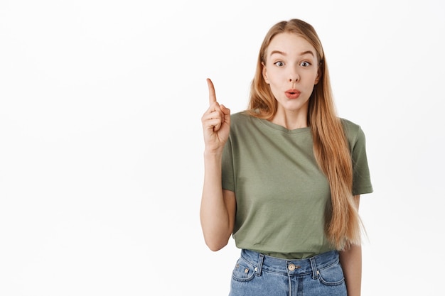 Eccitata ragazza bionda ansimando, dì wow, indicando la pubblicità del logo, mostrando il posto del banner per il tuo testo promozionale sopra, in piedi in una maglietta casual contro il muro bianco