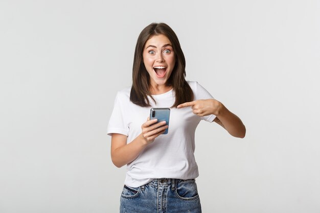 Eccitata ragazza bruna sorridente stupita e indicando lo schermo dello smartphone.