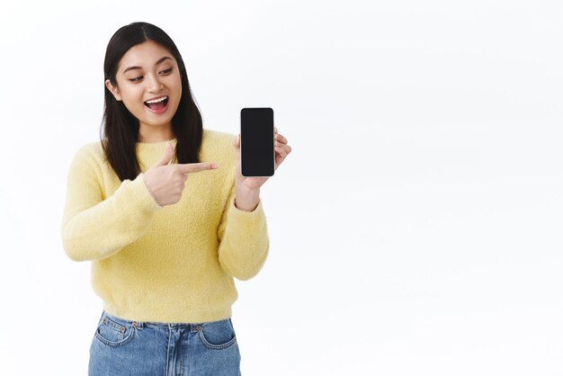 Eccitata ragazza carina che mostra il suo gioco preferito sullo schermo dello smartphone puntando e mostrando il display mobile con faccia allegra divertito sorridente ottimista discutere la fantastica applicazione sfondo bianco