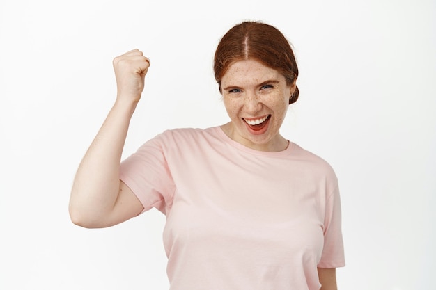 Eccitata ragazza dai capelli rossi, tifo per la squadra, pompa del pugno gioiosa e sorridente, determinata a vincere, tifare e cantare, vincere, celebrare la vittoria, in piedi sul bianco.