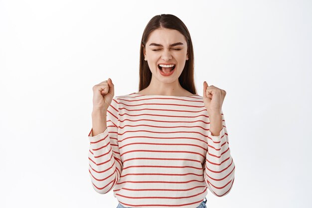 Eccitata ragazza di successo che celebra il successo, vince e grida sì, fa pompa a pugno, ottiene il suo obiettivo, trionfa della vittoria, in piedi sul muro bianco