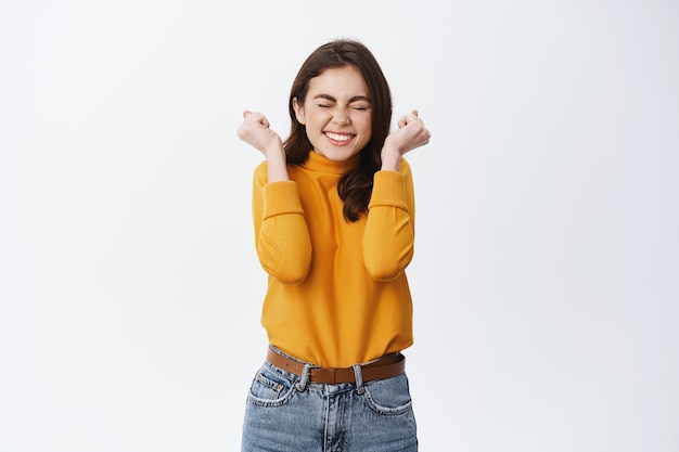 Eccitata ragazza sorridente che vince e celebra, stringe le mani allegre e salta dalla gioia, trionfa, raggiunge l'obiettivo o il premio, muro bianco