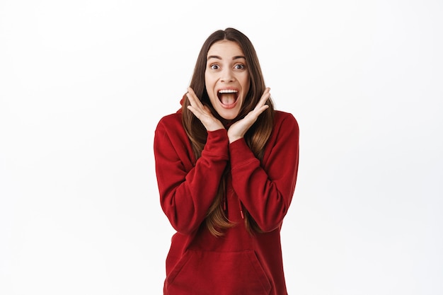 Eccitata ragazza vincente urla da gioiosa sorpresa fresca, bocca aperta affascinata, fissando impressionata e felice davanti, festeggiando, in piedi sul muro bianco