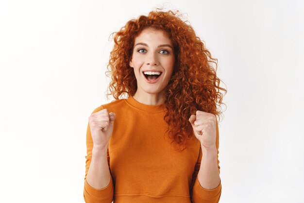 Eccitata splendida ragazza dai capelli rossi che fa il tifo per la squadra del cuore Ragazza riccia stringe i pugni motivata e ambiziosa sorridente ampiamente tifo trionfante guardando la partita di calcio celebrare la vittoria e il successo