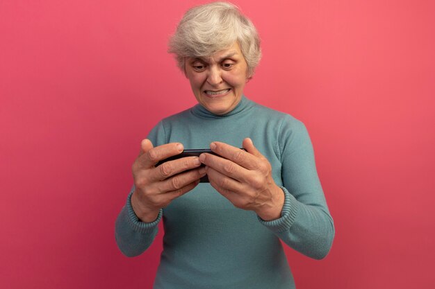 Eccitata vecchia donna che indossa un maglione a collo alto blu che gioca al telefono isolato sul muro rosa