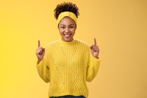 Eccitato affascinante giovane ragazza afro-americana acconciatura afro in fascia maglione che punta verso l'alto le dita indice sorridendo ridacchiando chiedendo di acquistare il fidanzato fantastico cosmetici b-day presente.