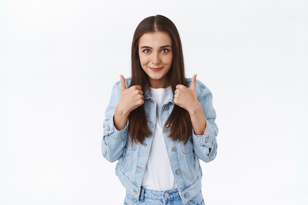 Eccitato, allegro, elegante, caucasico, femmina, bruna in giacca di jeans, mostrando il pollice in su e sorridendo felice, approva un'idea straordinaria e annuisce d'accordo, come una bella offerta, sfondo bianco