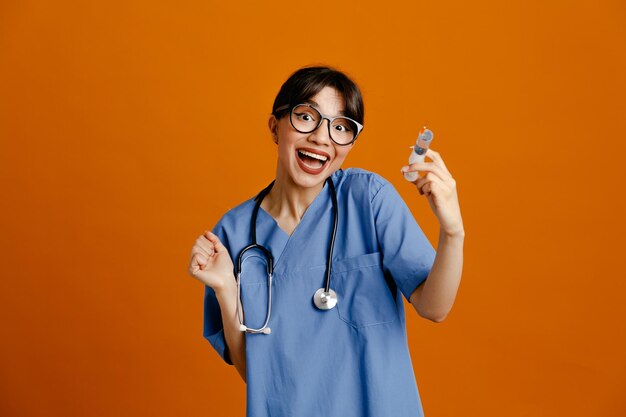 Eccitato azienda siringa giovane medico femminile che indossa uniforme fith stetoscopio isolato su sfondo arancione