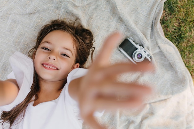 Eccitato bambino femminile con pelle leggermente abbronzata agghiacciante sulla coperta con le mani in alto e sorridente. Foto dall'alto all'aperto di una ragazza gioiosa in abito bianco sdraiata accanto alla telecamera sull'erba.