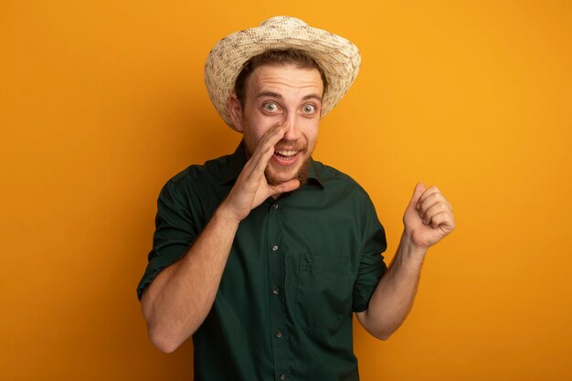 Eccitato bell'uomo biondo con cappello da spiaggia tiene la mano vicino alla bocca e tiene il pugno isolato sulla parete arancione