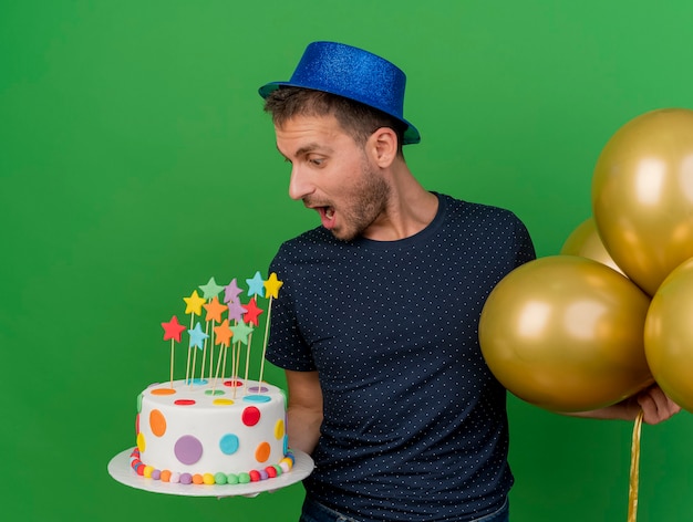 Eccitato bell'uomo caucasico che indossa il cappello blu del partito tiene palloncini di elio e guarda la torta di compleanno isolata su sfondo verde con spazio di copia