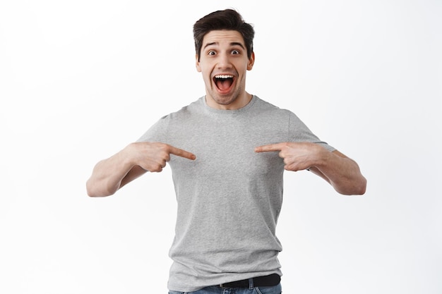 Eccitato bell'uomo urla di gioia, indicando al centro la t-shirt, mostrando testo promozionale e sorridendo stupito, in piedi su sfondo bianco