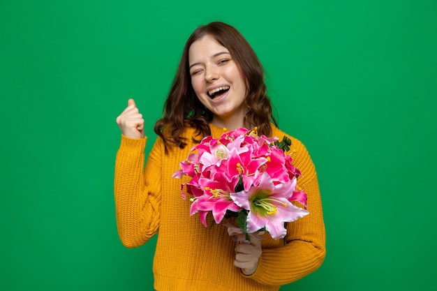 Eccitato che mostra sì gesto bella ragazza il giorno della donna felice che tiene il mazzo isolato sul muro verde