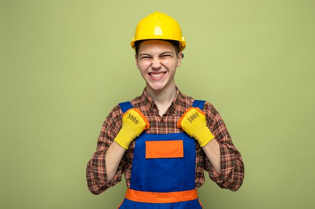 Eccitato che mostra sì gesto giovane costruttore maschio che indossa l'uniforme con i guanti