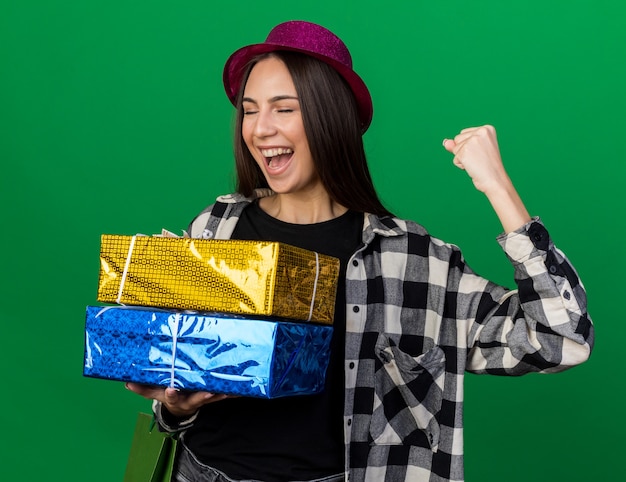 Eccitato con gli occhi chiusi giovane bella ragazza che indossa un cappello da festa che tiene scatole regalo con sacchetto regalo che mostra sì gesto isolato sul muro verde