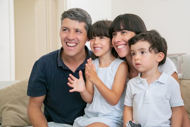 Eccitato coppia genitore gioioso con due bambini che guardano la TV, seduto sul divano nel soggiorno, distogliendo lo sguardo e sorridendo.