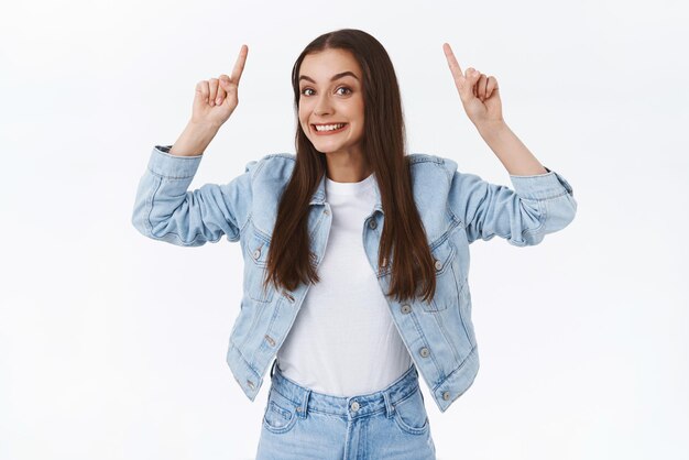 Eccitato divertente e bella bruna femmina caucasica in giacca di jeans jeans che punta il dito in alto sorridente allegro aspirante partecipare a un evento interessante consiglia di fare clic sul collegamento sfondo bianco