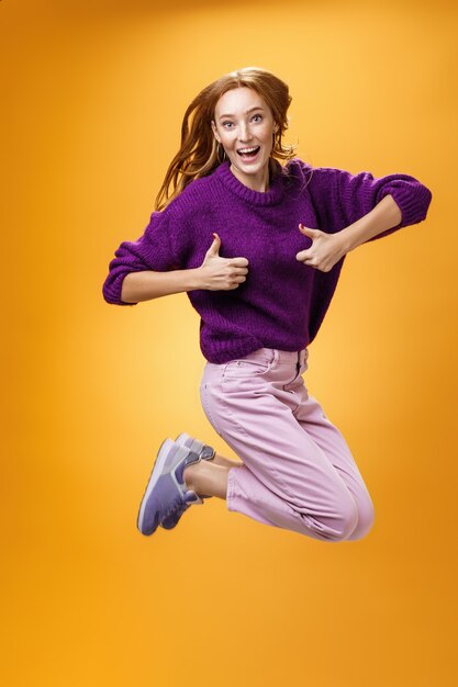 Eccitato e felice divertente giovane donna rossa in maglione viola che salta dalla felicità e dalla soddisfazione che mostra i pollici in su gesto di approvazione che dà risposta positiva e gradimento di vestiti fantastici