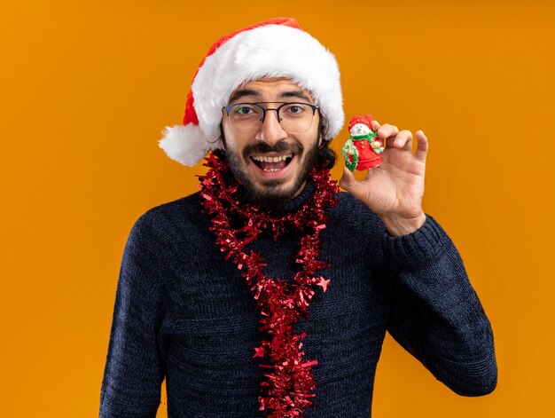 Eccitato giovane bel ragazzo che indossa il cappello di Natale con la ghirlanda sul collo tenendo il giocattolo isolato sulla parete arancione