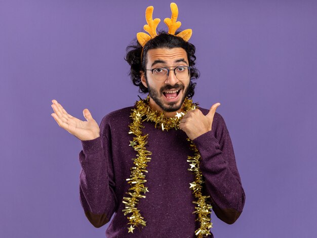 Eccitato giovane bel ragazzo che indossa il cerchio dei capelli di Natale con la ghirlanda sul collo che diffonde la mano e punti sul lato isolato sulla parete blu con lo spazio della copia