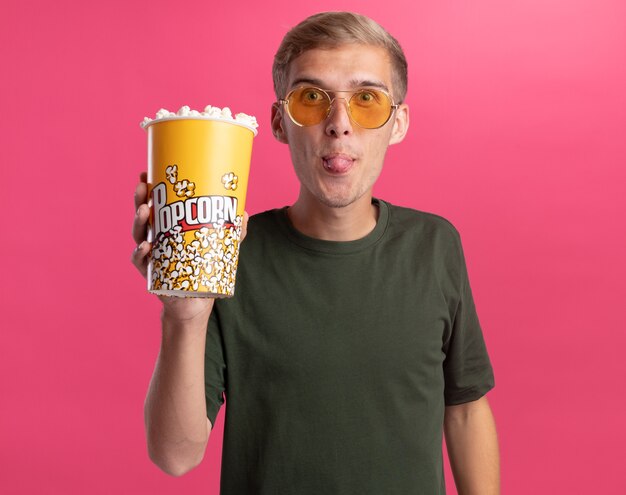 Eccitato giovane bel ragazzo che indossa la camicia verde e bicchieri tenendo il secchio di popcorn che mostra la lingua isolata sulla parete rosa