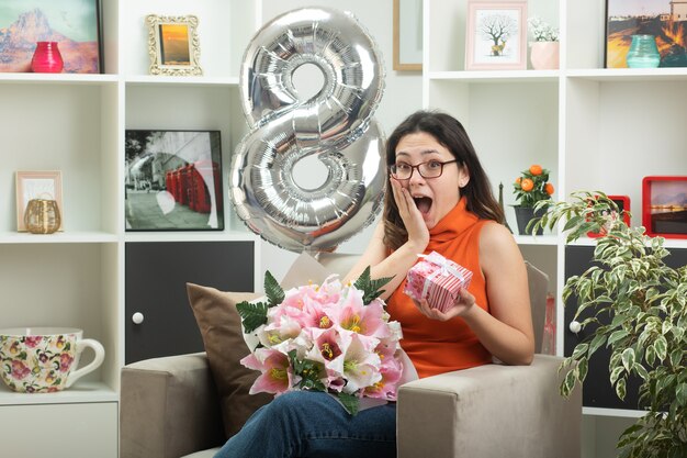 Eccitato giovane bella donna in bicchieri con bouquet di fiori e confezione regalo seduto sulla poltrona in soggiorno marzo giornata internazionale della donna