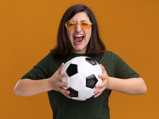 Eccitato giovane bella ragazza caucasica in occhiali da sole che tiene palla isolata sulla parete arancione con spazio di copia