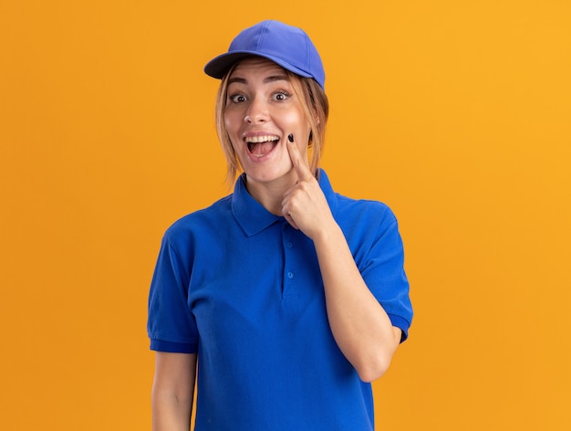 Eccitato giovane bella ragazza di consegna in uniforme mette il dito sul viso e guarda la telecamera sull'arancio