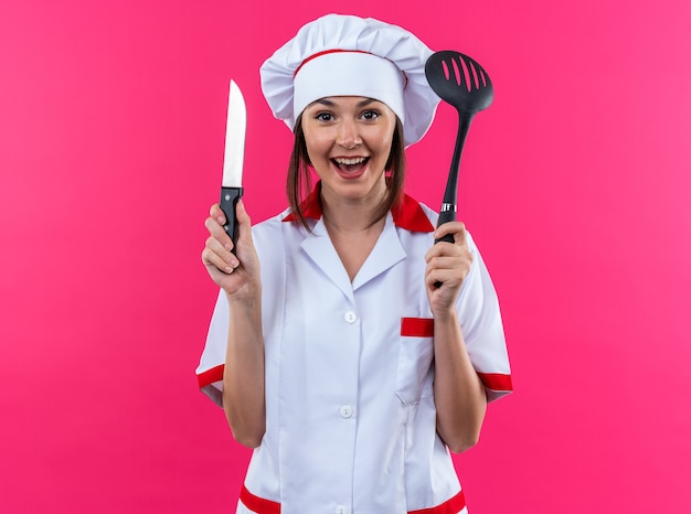 Eccitato giovane cuoca che indossa l'uniforme da chef tenendo il coltello con spatola isolato su sfondo rosa