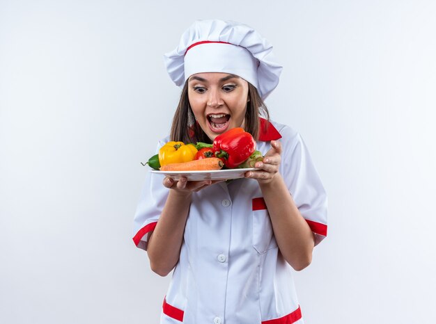 Eccitato giovane cuoca che indossa l'uniforme dello chef tenendo e guardando le verdure sul piatto isolato su parete bianca