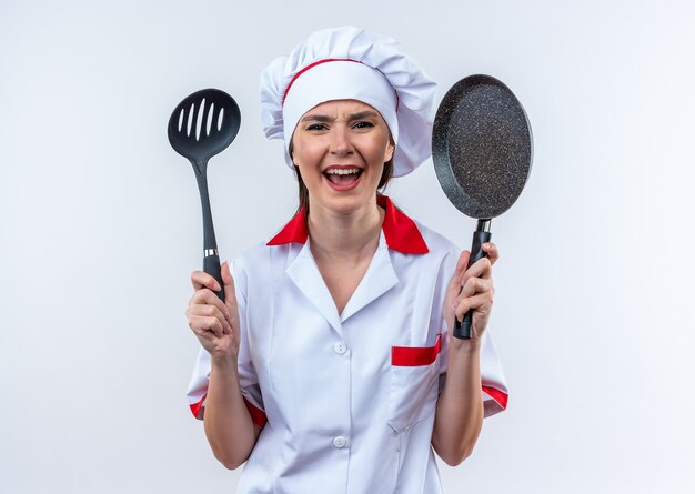 Eccitato giovane cuoca che indossa l'uniforme dello chef tenendo la spatola con padella isolata su sfondo bianco