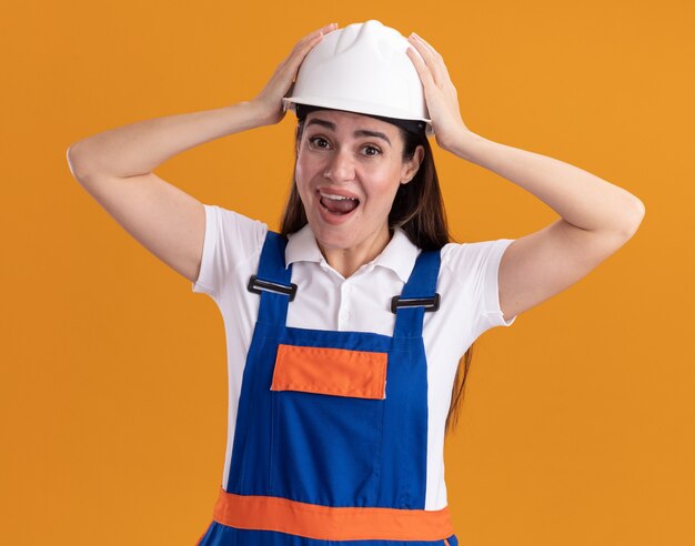 Eccitato giovane donna costruttore in uniforme ha afferrato la testa isolata sul muro arancione orange