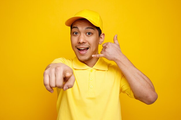 Eccitato giovane fattorino che indossa cappello e uniforme guardando e indicando la telecamera facendo gesto di chiamata