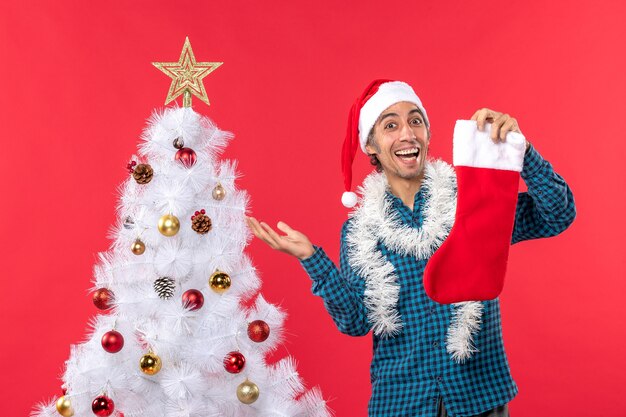 Eccitato giovane felice con il cappello di Babbo Natale in una camicia spogliata blu e tenendo il calzino di Natale vicino all'albero di Natale