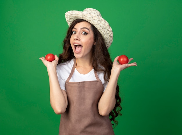 Eccitato giovane giardiniere femminile in uniforme che indossa cappello da giardinaggio tenendo i pomodori isolati sulla parete verde