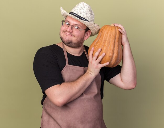 Eccitato giovane giardiniere maschio che indossa cappello da giardinaggio tenendo e guardando la zucca isolata sulla parete verde oliva