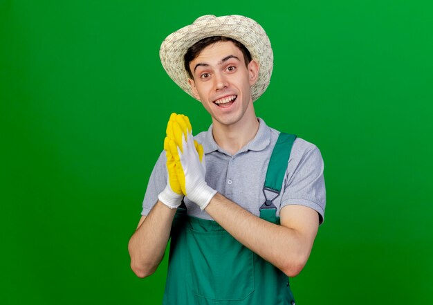 Eccitato giovane giardiniere maschio indossando guanti e cappello da giardinaggio tiene le mani insieme isolato su sfondo verde con spazio di copia