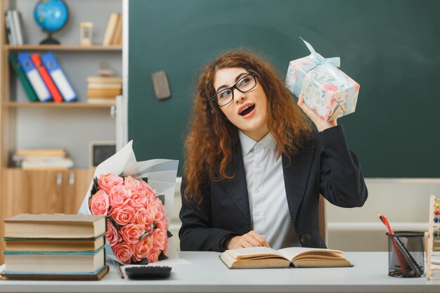 eccitato giovane insegnante femminile che tiene il regalo seduto alla scrivania con gli strumenti della scuola in classe