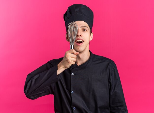 Eccitato giovane maschio biondo cuoco in uniforme da chef e berretto guardando la telecamera che copre metà del viso con frusta isolata sulla parete rosa