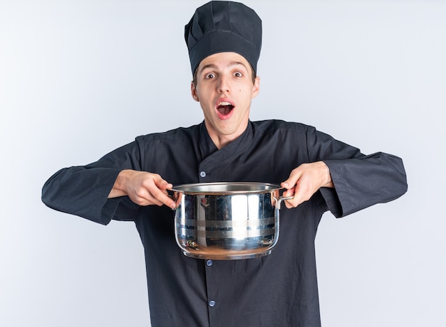 Eccitato giovane maschio biondo cuoco in uniforme da chef e cappello che tiene pentola con entrambe le mani