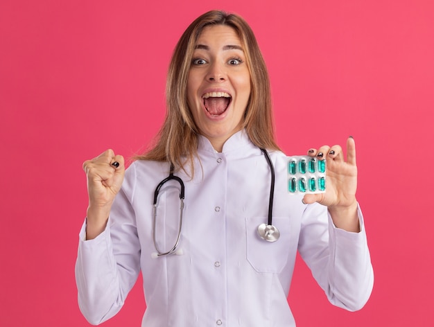 Eccitato giovane medico femminile che indossa abito medico con lo stetoscopio che tiene le pillole che mostrano sì gesto isolato sulla parete rosa