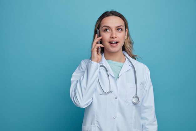 Eccitato giovane medico femminile che indossa abito medico e stetoscopio intorno al collo, alzando lo sguardo a parlare sul telefono