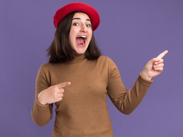 Eccitato giovane ragazza abbastanza caucasica con cappello berretto che punta a lato sulla porpora