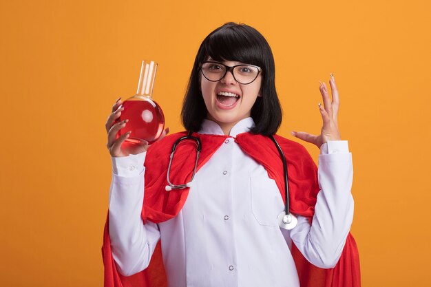 Eccitato giovane ragazza del supereroe che indossa uno stetoscopio con veste medica e mantello con gli occhiali che tengono la bottiglia di vetro chimica riempita con mano di diffusione del liquido rosso