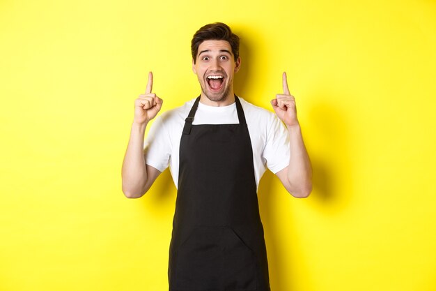 Eccitato proprietario di una caffetteria in grembiule nero che punta le dita in alto, mostrando offerte speciali, in piedi su sfondo giallo