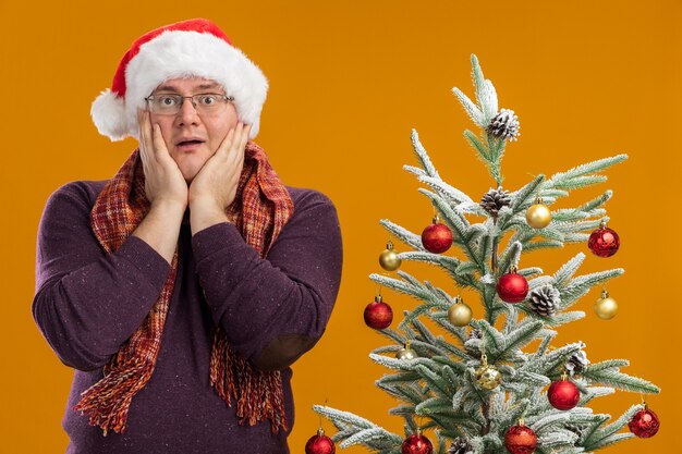 eccitato uomo adulto con gli occhiali e cappello da Babbo Natale con sciarpa intorno al collo in piedi vicino all'albero di Natale decorato tenendo le mani sul viso guardando la telecamera isolata su sfondo arancione