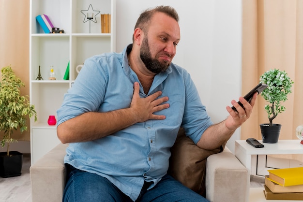 Eccitato uomo adulto slavo si siede sulla poltrona mettendo la mano sul petto guardando il telefono all'interno del soggiorno