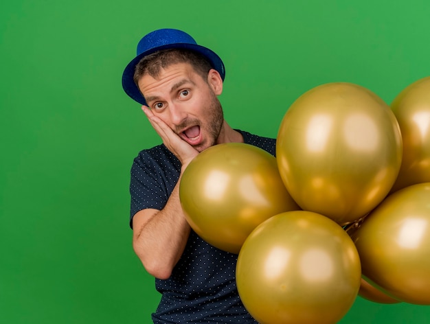 Eccitato uomo bello che indossa il cappello da festa blu mette la mano sul viso e tiene palloncini di elio isolati sulla parete verde