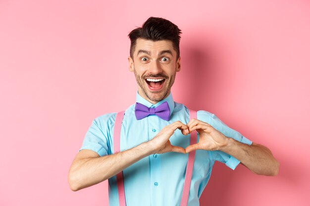 Eccitato uomo sorridente che mostra il cuore che batte e guarda con amore, in piedi su un romantico sfondo rosa. Il giorno di San Valentino concetto.