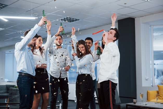 Ecco come appare il successo. Foto di giovane squadra in abiti classici che celebra il successo mentre si tiene un drink nel moderno ufficio illuminato